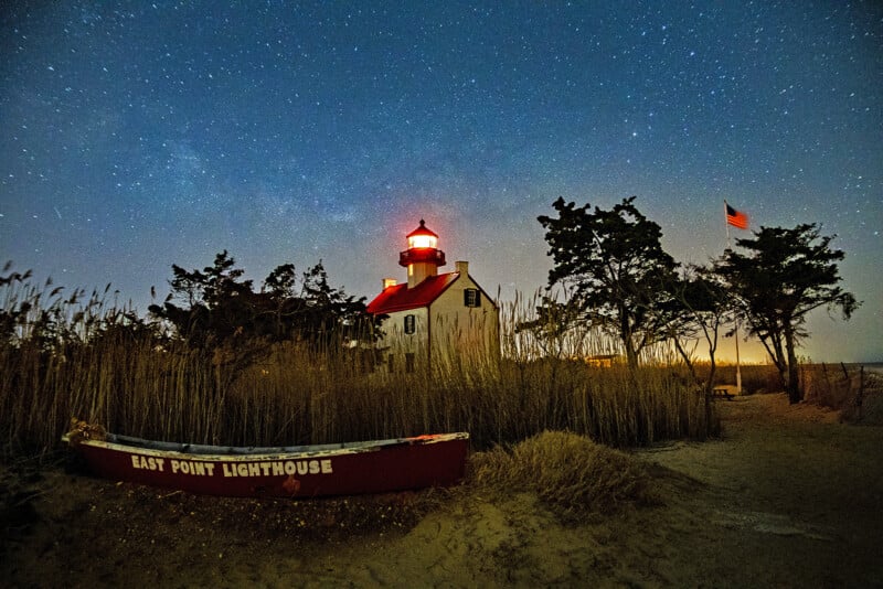 A red-roofed lighthouse glows under a starry night sky, surrounded by tall grass and trees. An American flag waves nearby, and a red boat labeled "East Point Lighthouse" rests in the foreground.