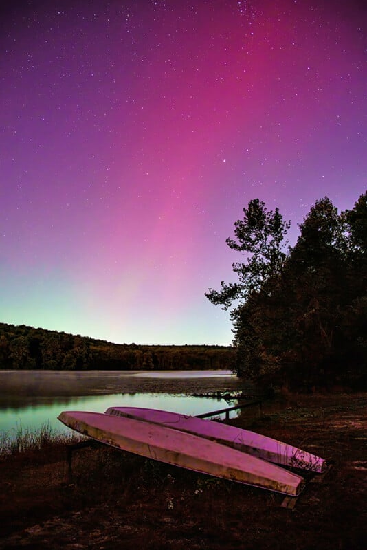 A vibrant night sky glows pink and purple above a calm lake, with two upside-down boats resting on the grassy shore beside trees and distant hills.