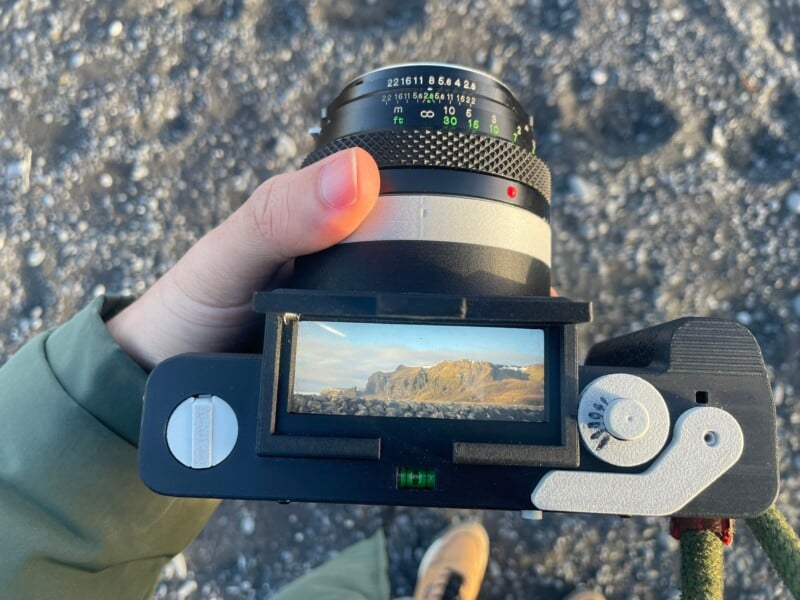 A person holding a camera, focusing on a landscape visible in the viewfinder. The camera screen displays mountains and a blue sky, while the ground below is rocky and the person's shoes are partially visible.