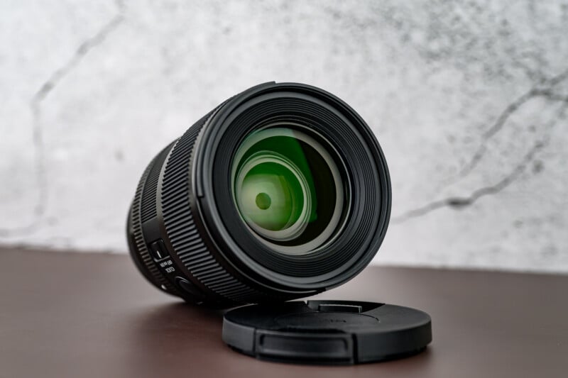 A black camera lens with a visible green reflection is placed on its side on a brown surface, next to a detached lens cap, with a textured gray and white background.