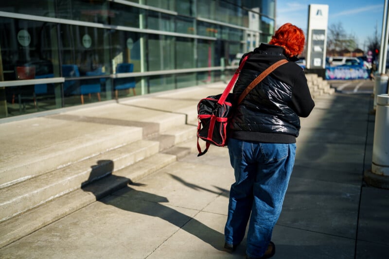 A person with bright red hair, wearing a black jacket and blue jeans, walks on a sunny sidewalk next to a modern glass building, carrying a black and pink bag over their shoulder.
