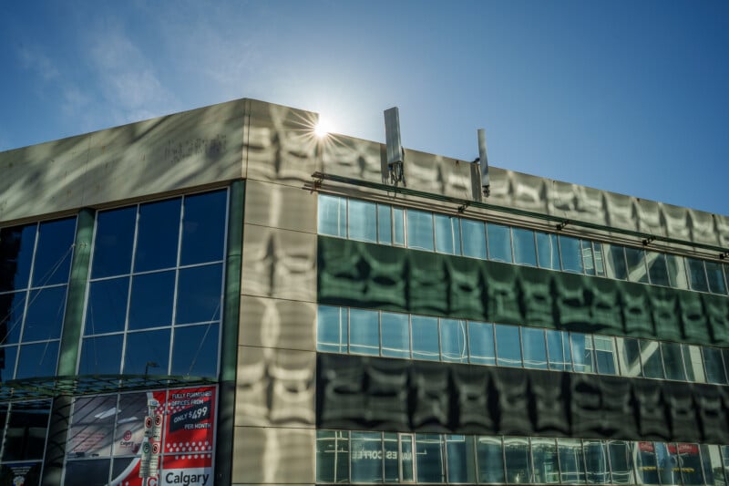 A modern office building with glass windows reflects sunlight, creating wavy light patterns on its facade. The sun is partially visible behind the rooftop, and a blue sky is in the background.