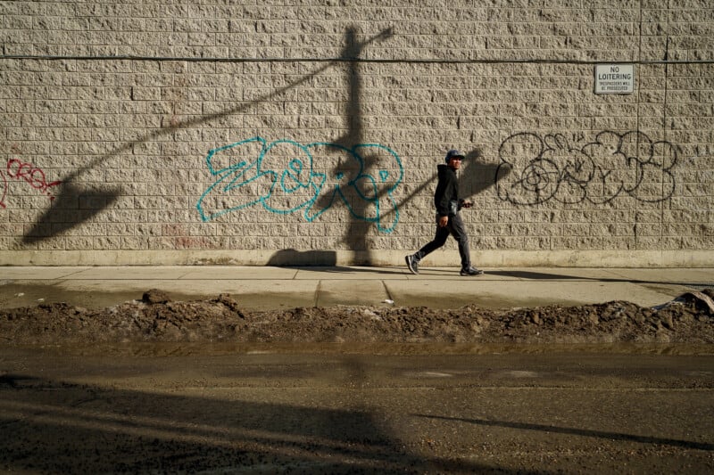A person in dark clothing walks along a sidewalk beside a brick wall with graffiti. The wall casts a shadow from a streetlight, and a "No Loitering" sign is visible. The street is muddy with puddles in the foreground.