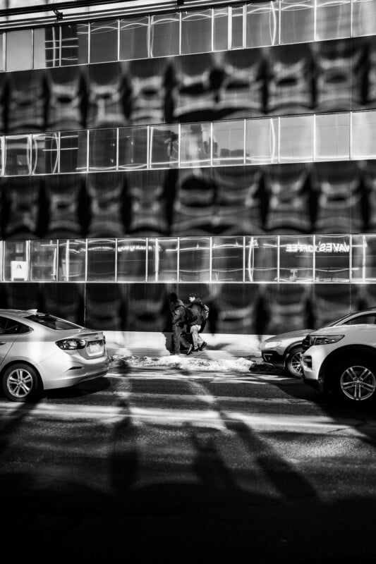 Two people stand and talk on a snowy sidewalk between parked cars, with dramatic shadow patterns cast on the street and building behind them; scene is in black and white.