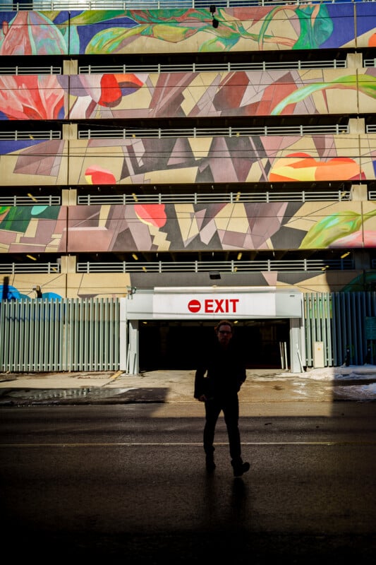 A person in silhouette walks in front of a parking garage exit. The garage exterior features a colorful, abstract mural with leaves and geometric shapes. A bright EXIT sign is visible above the entrance.