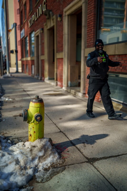 A yellow fire hydrant surrounded by snow is on a city sidewalk. In the background, a security guard in black uniform walks past a red brick building.