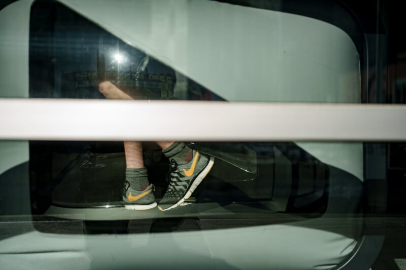 A person’s legs wearing gray socks and black-and-orange Nike sneakers are visible through the glass of a modern building or vehicle, with sunlight reflecting on the glass.