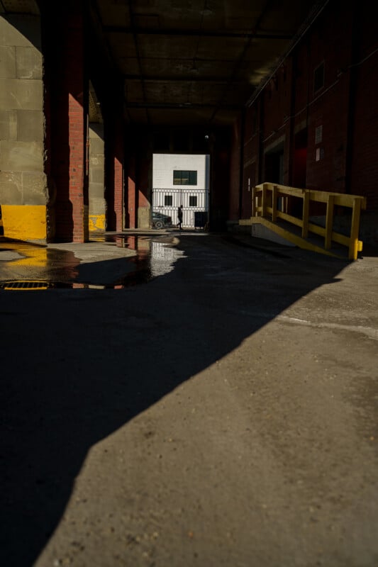 Shadowed alleyway with concrete and brick walls, a yellow railing on the right, and sunlight illuminating part of the ground. A small puddle reflects light near the alley’s entrance, with a white building visible in the background.