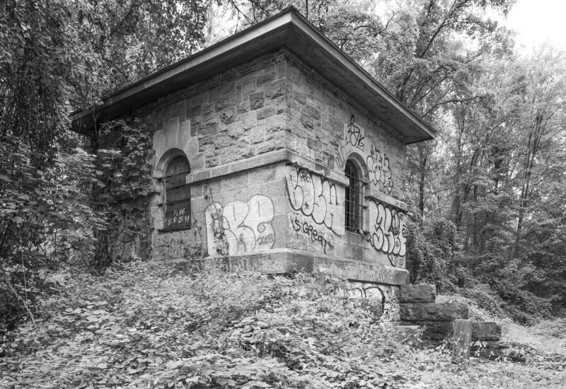A small, old stone building covered in graffiti stands surrounded by dense, overgrown vegetation and trees. The structure has arched windows and appears weathered, blending into the forested background.