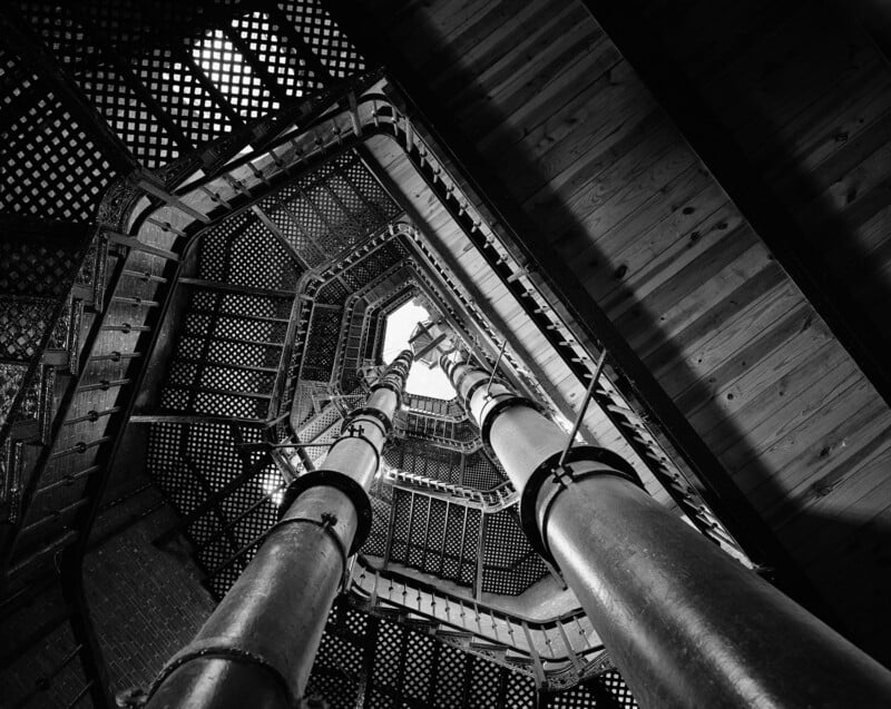Black and white photo looking up the center of a metal spiral staircase with three large vertical pipes, geometric patterns, and a view of the sky framed by the upper opening.
