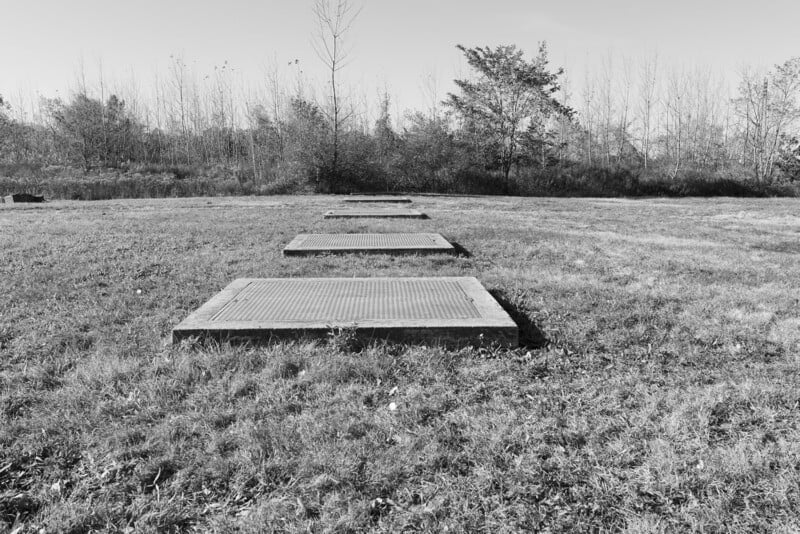 Four square concrete slabs are arranged in a row on a grassy field, with trees and bushes in the background under a clear sky. The image is in black and white.