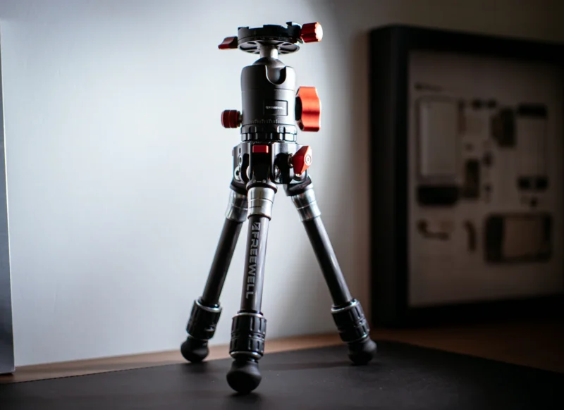 A compact camera tripod with metallic legs and red adjustment knobs stands on a desk, illuminated by natural light. A framed technical diagram is visible in the blurred background.