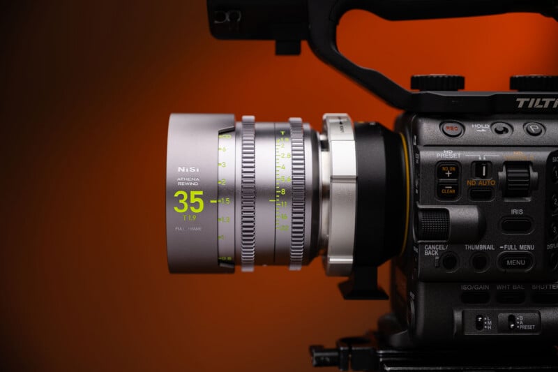 Close-up of a professional video camera with a NiSi 35mm cine lens attached, showing detailed focus and aperture markings, against a warm orange background.