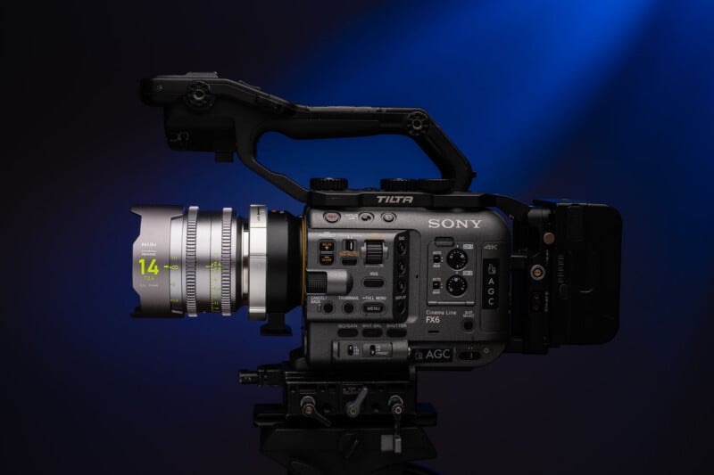 A professional Sony video camera with various control buttons and a Tilta rig attachment is mounted on a tripod against a dark background with a blue spotlight.