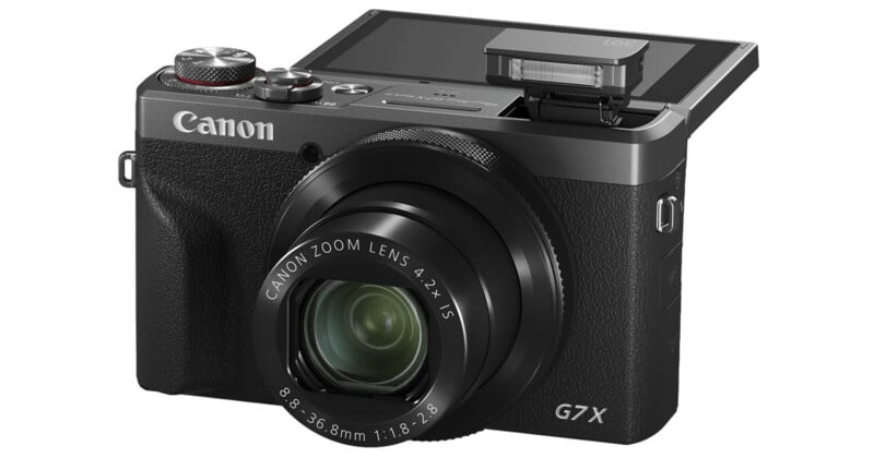 A Canon G7 X digital camera with its LCD screen flipped up and the built-in flash raised, showing the lens and controls on the front and top of the camera.