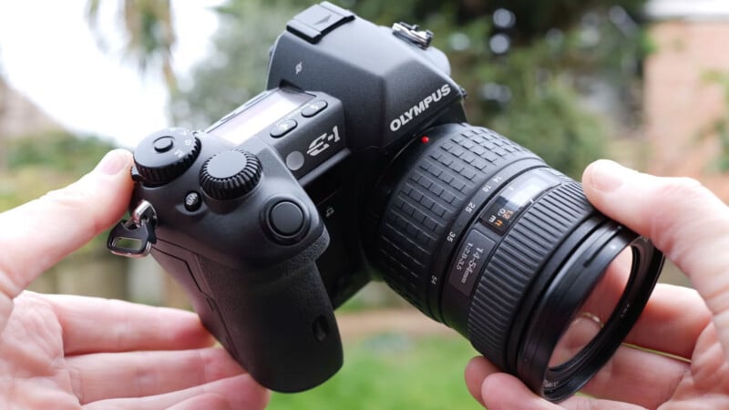 A person holds a black Olympus E-1 digital camera with a large lens outdoors, with a blurred green and brown background.