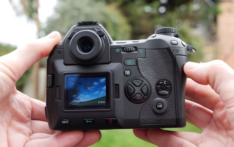 A person holds a digital camera outdoors, showing the camera’s back with a screen displaying a photo of tree branches and a blue sky. Control buttons and dials are visible around the screen.