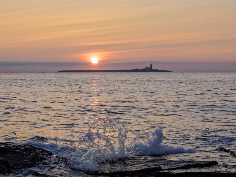 Sunset over the sea with waves splashing against rocks in the foreground and a distant silhouette of an island with a lighthouse on the horizon. The sky is tinged with warm orange and pink hues.