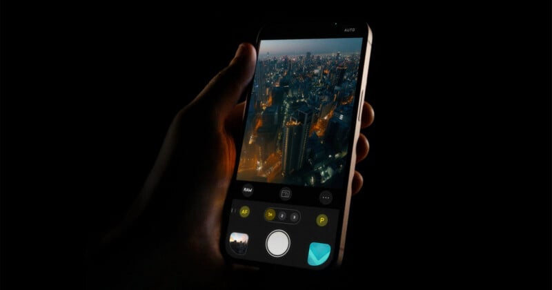A hand holds a smartphone in the dark, displaying a cityscape photo on the screen with camera app controls visible, capturing a night view of tall, illuminated buildings.