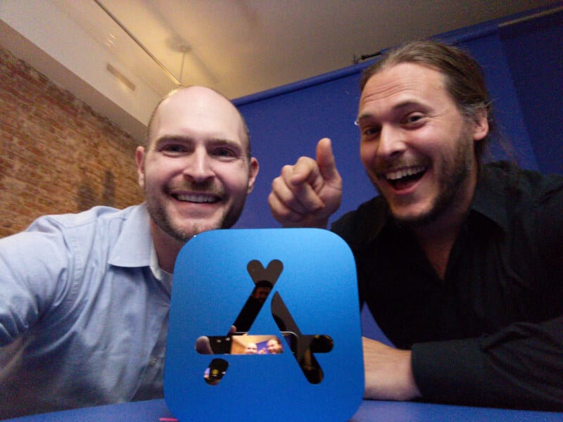Two smiling men pose for a selfie behind a blue Apple App Store award. One man points at the award, and their reflection is visible on its surface. They are indoors with a brick wall in the background.
