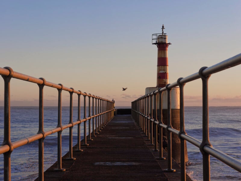 A long pier with metal railings leads to a red and white striped lighthouse by the sea at sunset. A bird is flying near the lighthouse and the sky is clear with soft pastel colors.