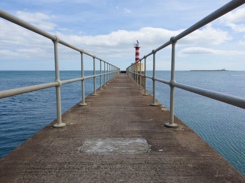 A concrete pier with metal railings extends over calm blue water toward a red and white striped lighthouse under a partly cloudy sky. An island is visible in the distance on the right.