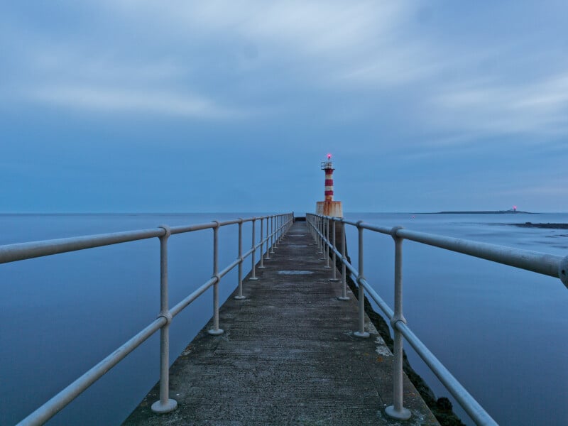 A concrete pier with metal railings leads to a red and white striped lighthouse, standing over calm blue water under a cloudy sky at dusk.