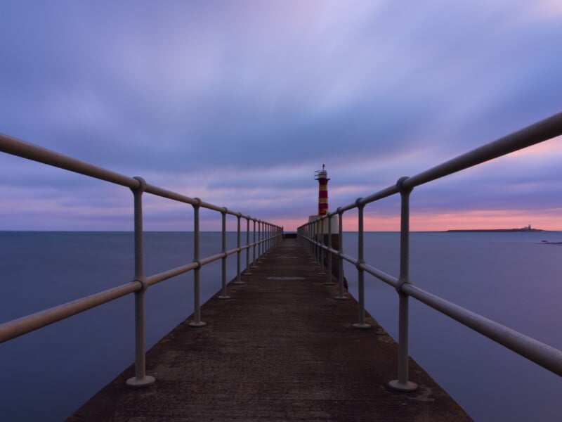A long pier with metal railings extends over calm water toward a red and yellow striped lighthouse at sunset, with colorful clouds and a distant shoreline in the background.