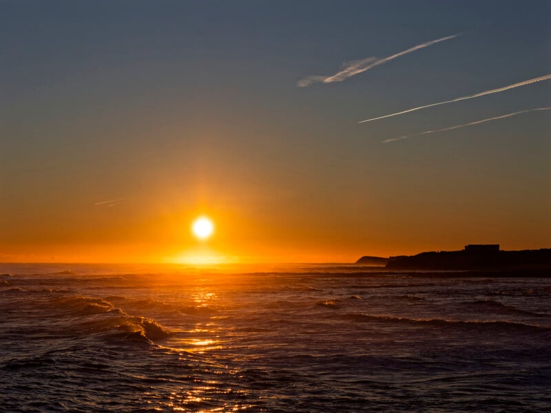 A vibrant orange sunset over the ocean, with the sun low on the horizon, gentle waves reflecting the sunlight, and silhouettes of distant cliffs beneath a clear sky with a few clouds and contrails.