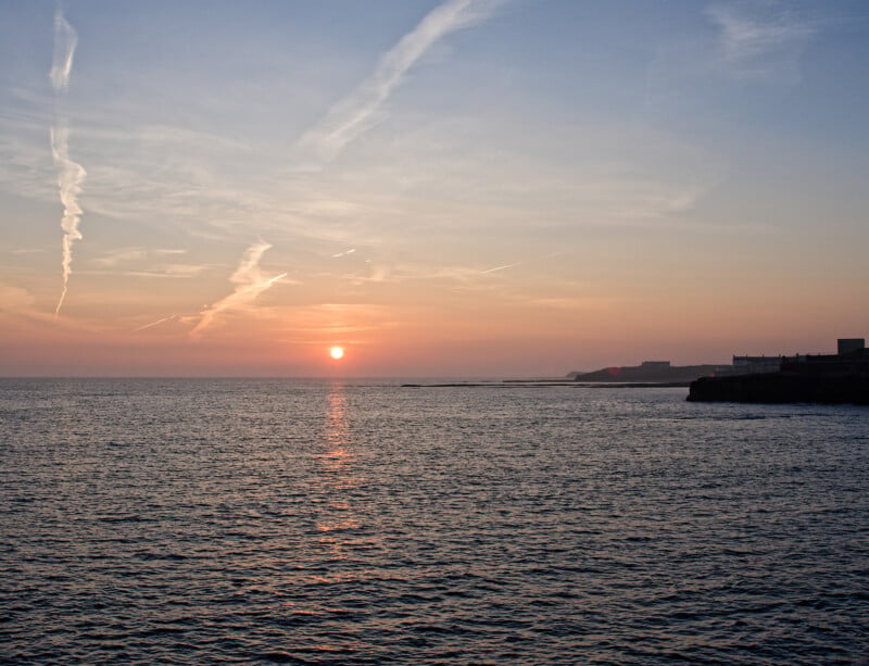 A serene sunset over a calm sea, with the sun low on the horizon, casting a warm reflection on the water. Wispy clouds streak across the sky and dark silhouettes of distant land appear on the right.