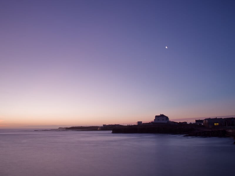 A calm coastal scene at dusk with soft purple and blue tones in the sky, a crescent moon above, and a house and buildings silhouetted against the horizon near the water’s edge.