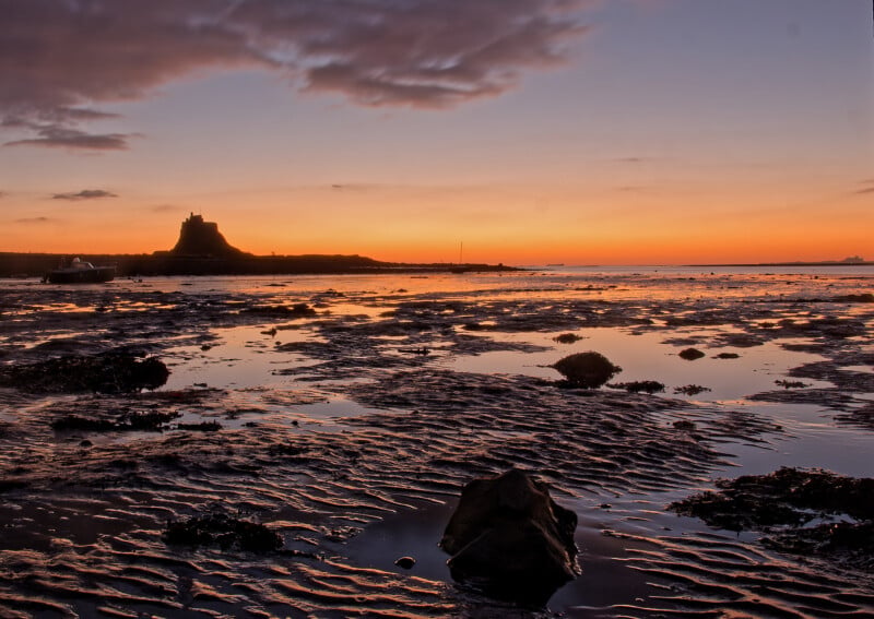 A dramatic coastal landscape at sunset, with rippled wet sand and shallow pools reflecting orange and purple sky. A silhouetted island with a castle sits in the distance, under scattered clouds.