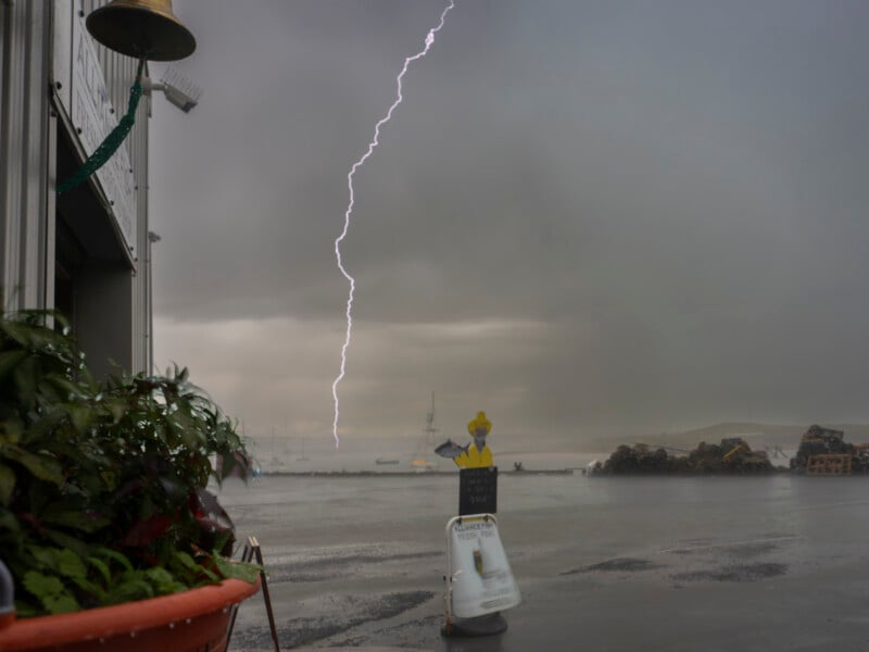 A bolt of lightning strikes over a foggy harbor, with boats in the distance. In the foreground, a plant pot and a yellow safety sign shaped like a person stand on the wet ground near a building.