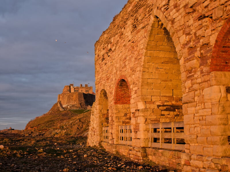 A stone wall with large arches is lit by golden sunlight in the foreground, while a historic castle stands on a hill in the background under a cloudy sky. Rocks and pebbles cover the ground.