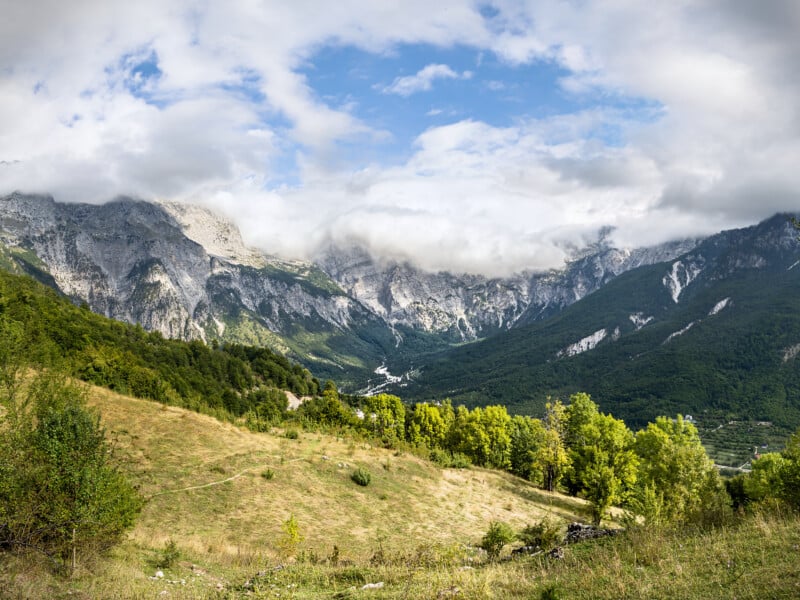 A scenic landscape of green hills and trees in the foreground, with rocky mountains partially covered by clouds and blue sky in the background.