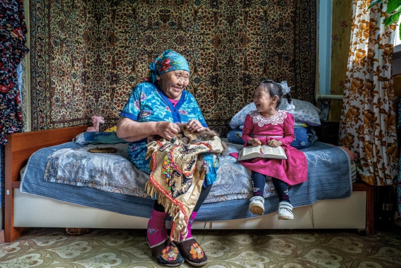 An elderly woman and a young girl sit on a bed in a cozy room. The woman embroiders fabric while the smiling girl in a red dress looks at her with a book in her lap. A patterned carpet hangs on the wall behind them.