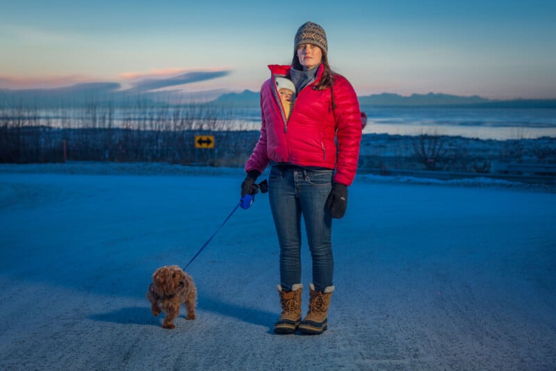 A woman in a red jacket, winter hat, and boots stands on a snowy road holding a small dog on a leash, with a baby in a carrier on her chest. Snowy landscape and sunset sky are in the background.