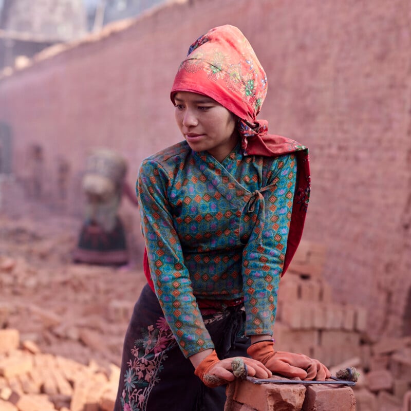 A woman wearing a patterned headscarf, long-sleeve shirt, and gloves stands among stacked bricks in front of a brick wall, looking to her left. The scene appears dusty and she is leaning on the bricks.