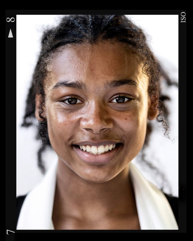 A close-up of a smiling young person with curly hair, wearing a black and white collared shirt, shown within a black rectangular frame with numbers and symbols on the edges.