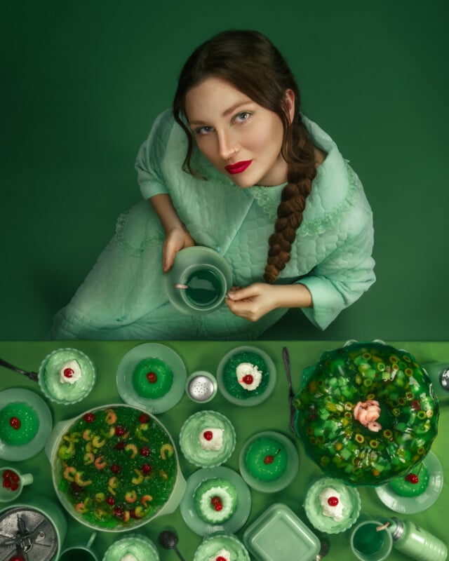A woman in a mint green outfit with a braid looks up while holding a green teacup. She is surrounded by vintage green dishes and various green foods on a matching green table and background.