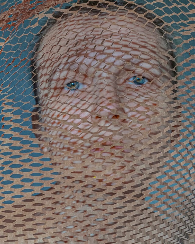 A person’s face seen through a sheet of textured, honeycomb-patterned material, which distorts their features. The background is blue, and the eyes, nose, and mouth appear blurred behind the mesh.