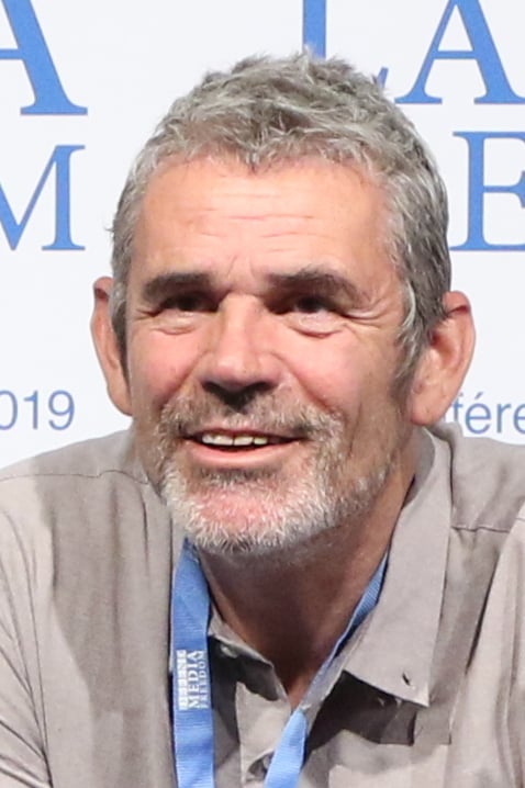 A middle-aged man with short gray hair and a salt-and-pepper beard smiles, wearing a gray shirt and a blue lanyard, sitting in front of a white backdrop with blue text.