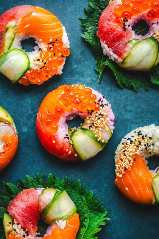 Colorful sushi donuts topped with slices of salmon, tuna, cucumber, sesame seeds, and orange fish roe, arranged on a dark blue surface with green leaves underneath some of the sushi pieces.