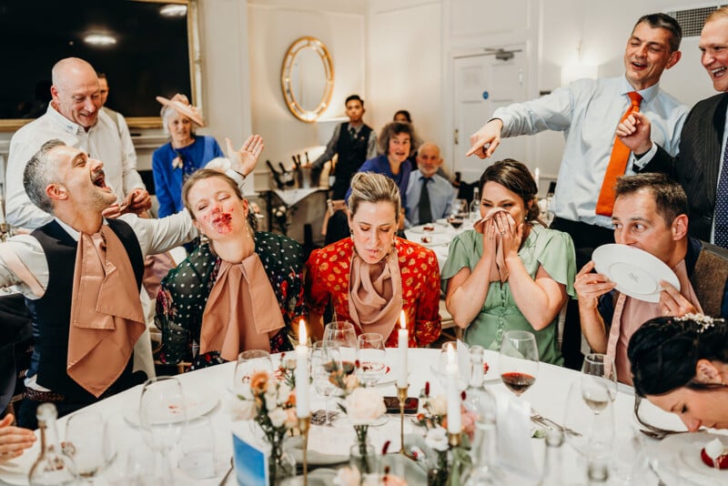 A group of well-dressed people sit around a table at a lively event. Some have cream or dessert on their faces and are laughing, while others cover their faces in amusement. The atmosphere is joyful and festive.