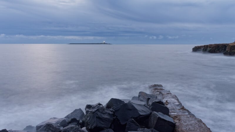 Large dark rocks and a stone jetty jut into a calm, misty sea under a cloudy blue-gray sky. In the distance, an island with a small lighthouse and a shining beacon is visible. The coastline is visible on the right.