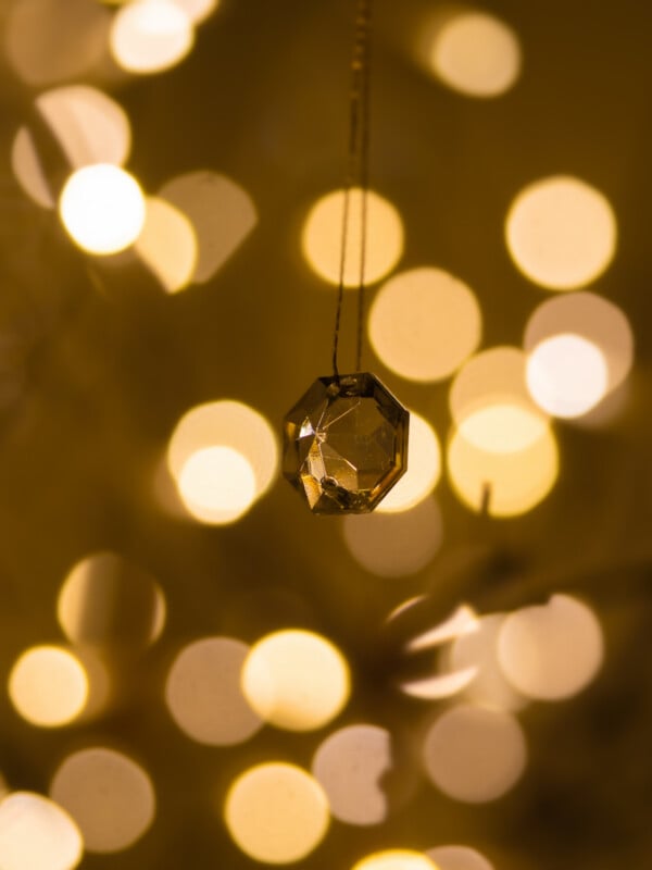 A faceted crystal ornament hangs from a thin string, with warm, golden bokeh lights softly blurred in the background, creating a festive, dreamy atmosphere.