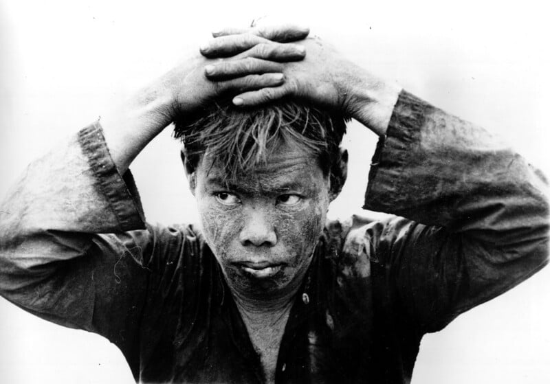 A person with a dirt-covered face and hands rests their hands on top of their head, looking to the side with a serious expression. They wear a weathered long-sleeve shirt. The image is in black and white.
