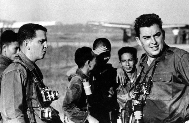 A group of photojournalists and local men stand outdoors, some with cameras hanging from their necks. One man shields his eyes from the sun as others look in different directions, appearing focused and alert.