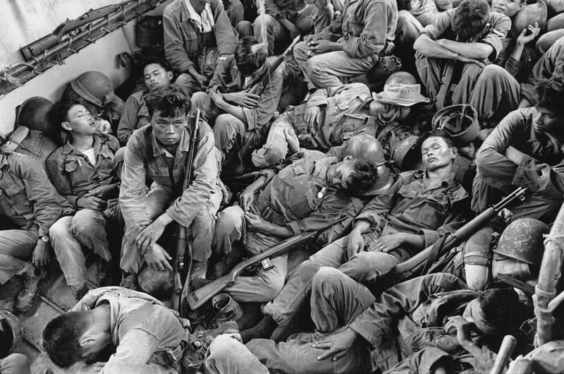 A group of weary soldiers in uniform, some sleeping and others resting, sit closely packed together on the floor of a military vehicle or boat, holding their rifles and gear.
