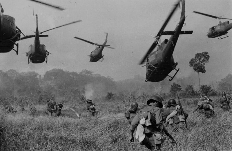 U.S. soldiers advance through tall grass as several helicopters hover overhead, preparing to land in a jungle clearing during a military operation. The scene is tense and filled with movement.